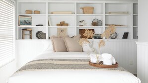 A neatly organized bedroom with a large bed, a built-in bookshelf, and a window with blinds. - Pier on Main (Tiverton)