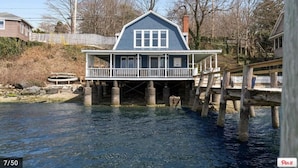 A house with a blue exterior and a wooden deck extending over a body of water. - Pier on Main (Tiverton)