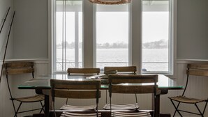 A dining area with a glass table and chairs, a wicker pendant light, and a view of a body of water through large windows. - Pier on Main (Tiverton)