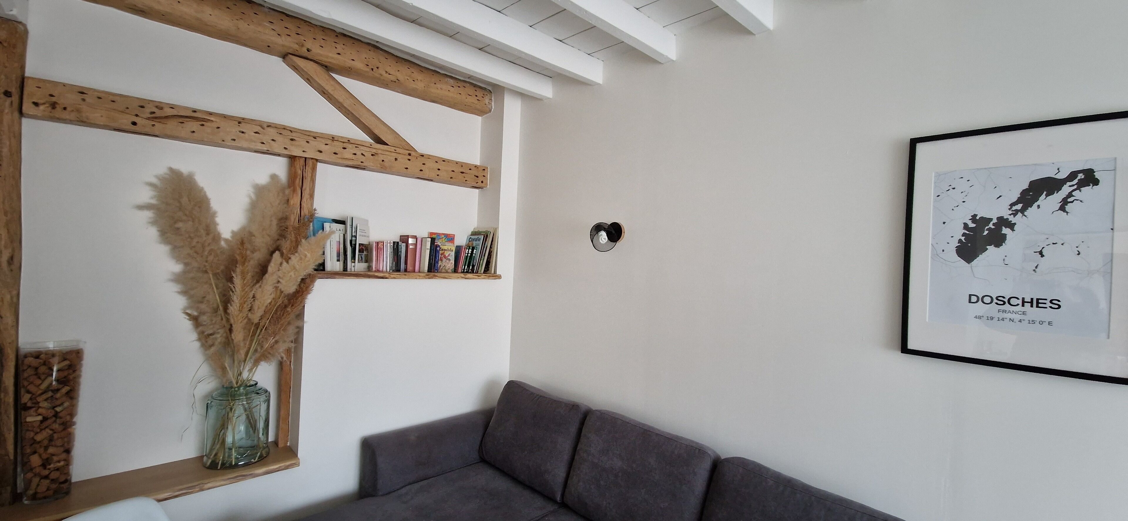 Una stanza con un divano grigio, uno scaffale in legno con libri e piante essiccate, e una mappa incorniciata appesa al muro.
