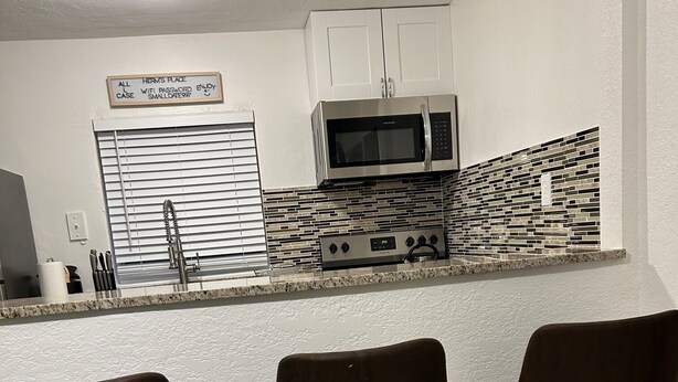 A kitchen with white cabinets, a microwave, a tiled backsplash, and three bar stools. - 7 mins to the beach (Delray Beach)