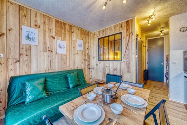 Une pièce aux murs lambrissés, avec un canapé vert, une table à manger dressée avec des assiettes et des verres, et une fenêtre donnant sur un beau paysage.