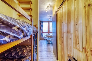 Um corredor estreito com paredes de madeira, uma beliche com roupa de cama estampada, um espaço de refeições com mesa e cadeiras, e uma janela com cortinas.