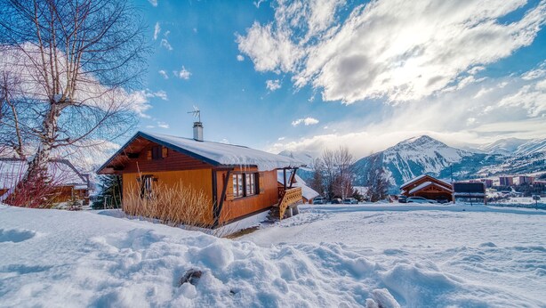 A wooden cabin with a snow-covered roof, a wooden deck, and a mountain backdrop. - Chalet les Colchiques (Fontcouverte-la-Toussuire)