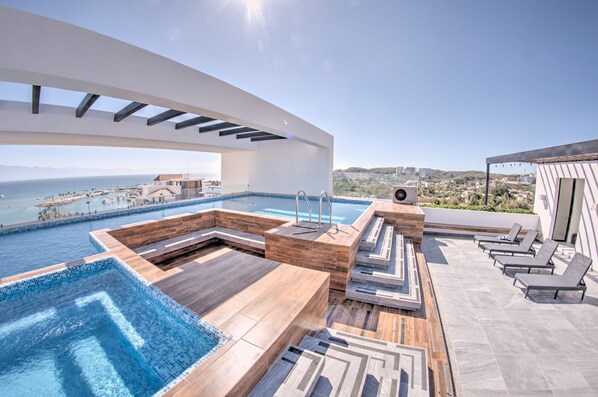 A rooftop infinity pool with wooden deck and lounge chairs, overlooking a coastal cityscape.