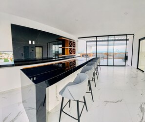 A modern kitchen with a black countertop, white marble backsplash, and a bar area with white bar stools. - Cruz 504: Studio w Rooftop Pool, 2 Blocks to Beach (Cruz de Huanacaxtle)