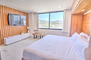 A hotel room with a large bed, a TV mounted on the wall, a window with a mountain view, and a wooden panel wall. - Cruz 504: Studio w Rooftop Pool, 2 Blocks to Beach (Cruz de Huanacaxtle)