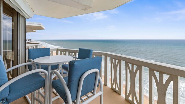 Un balcon avec vue sur l’océan, des balustrades blanches et des chaises bleues avec des coussins.