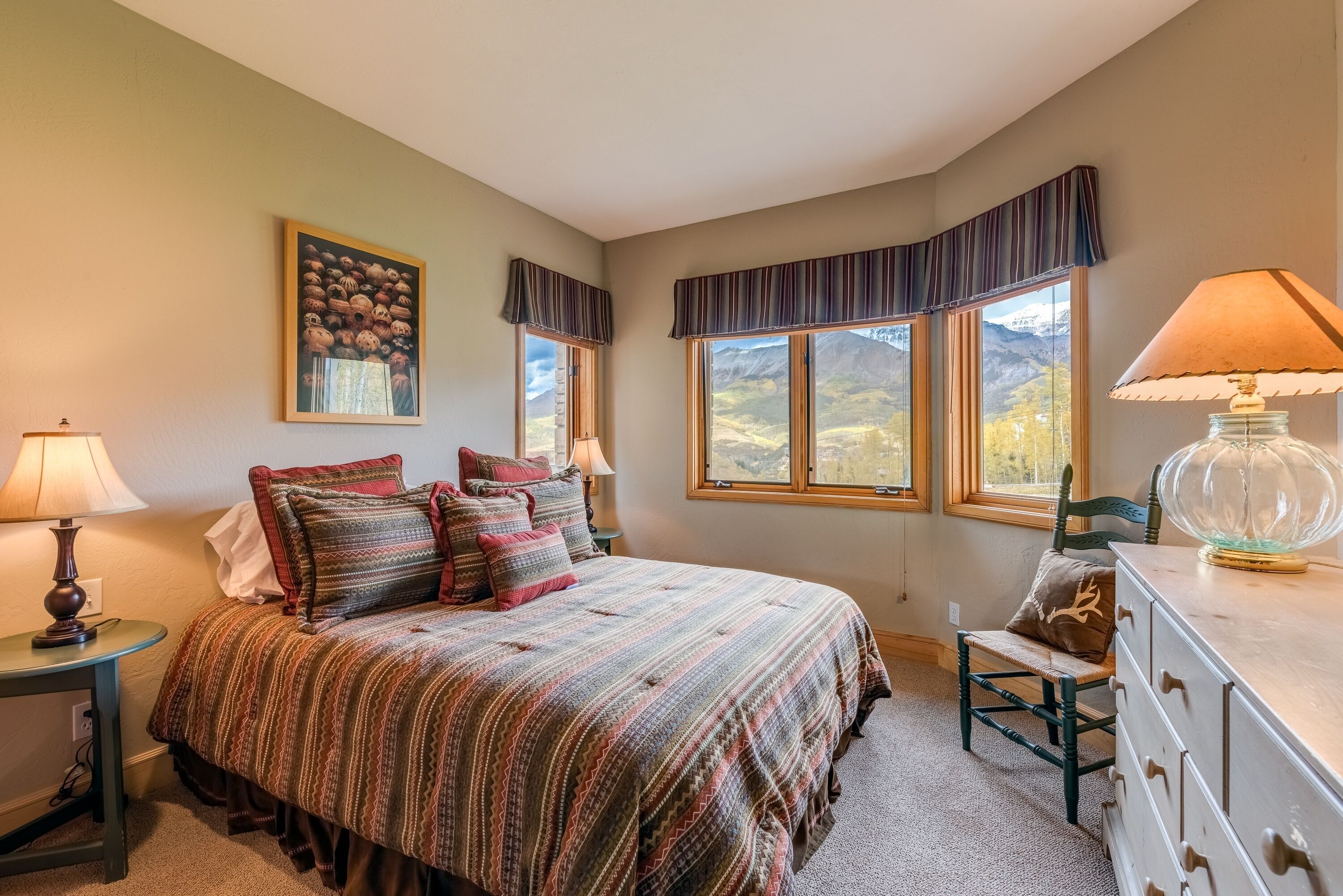 A bedroom with a large bed, two bedside lamps, a dresser, and a chair. There are windows with curtains and a view of mountains.