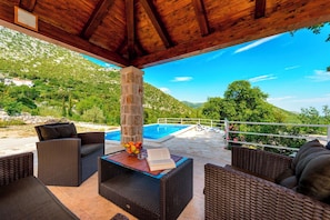 An outdoor seating area with wicker furniture, a stone pillar, and a view of a swimming pool and mountains.