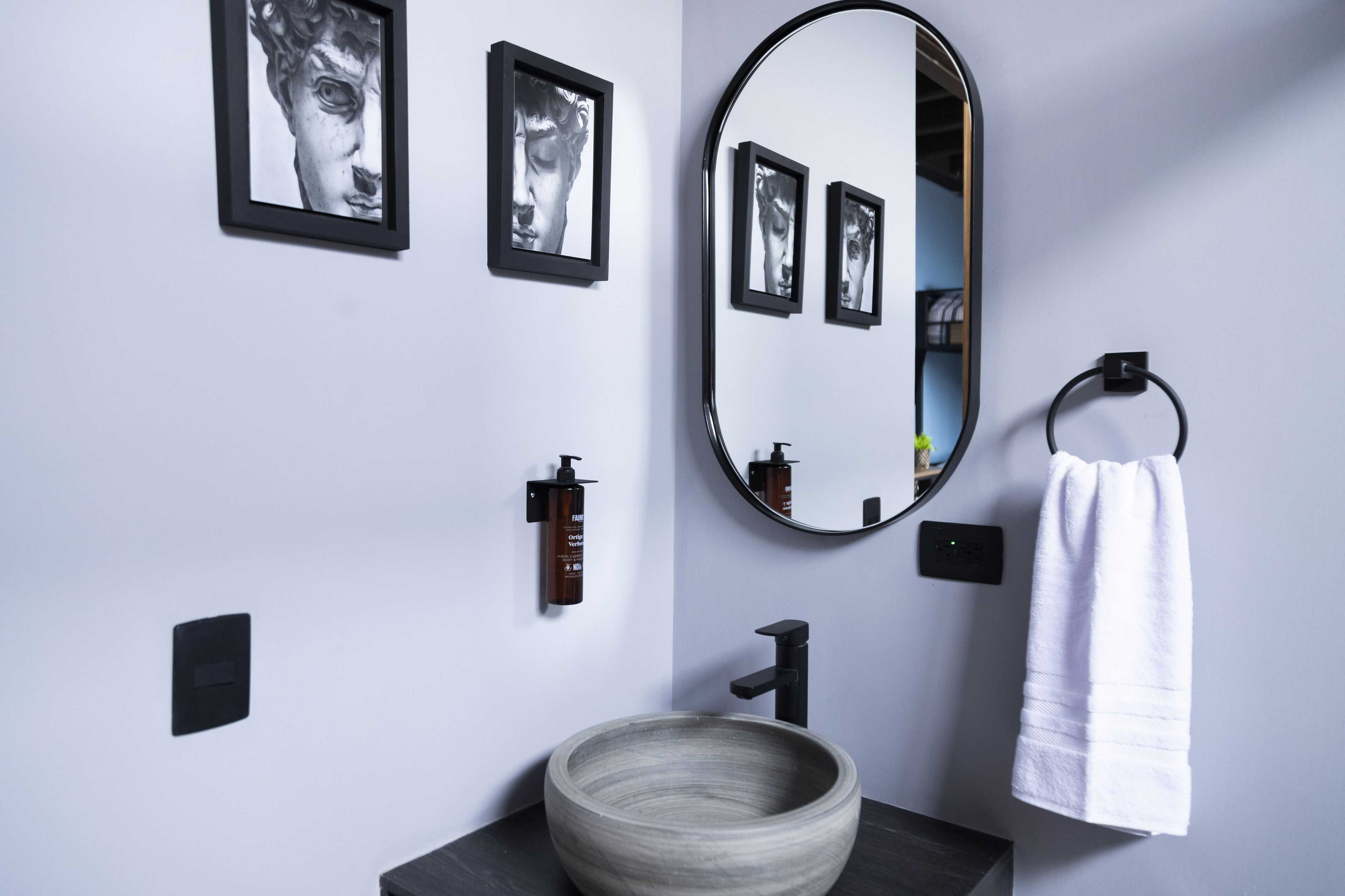 Une salle de bain avec un miroir rond, des tableaux encadrés de noir, une serviette blanche, un lavabo en forme de bol en bois et un robinet noir.