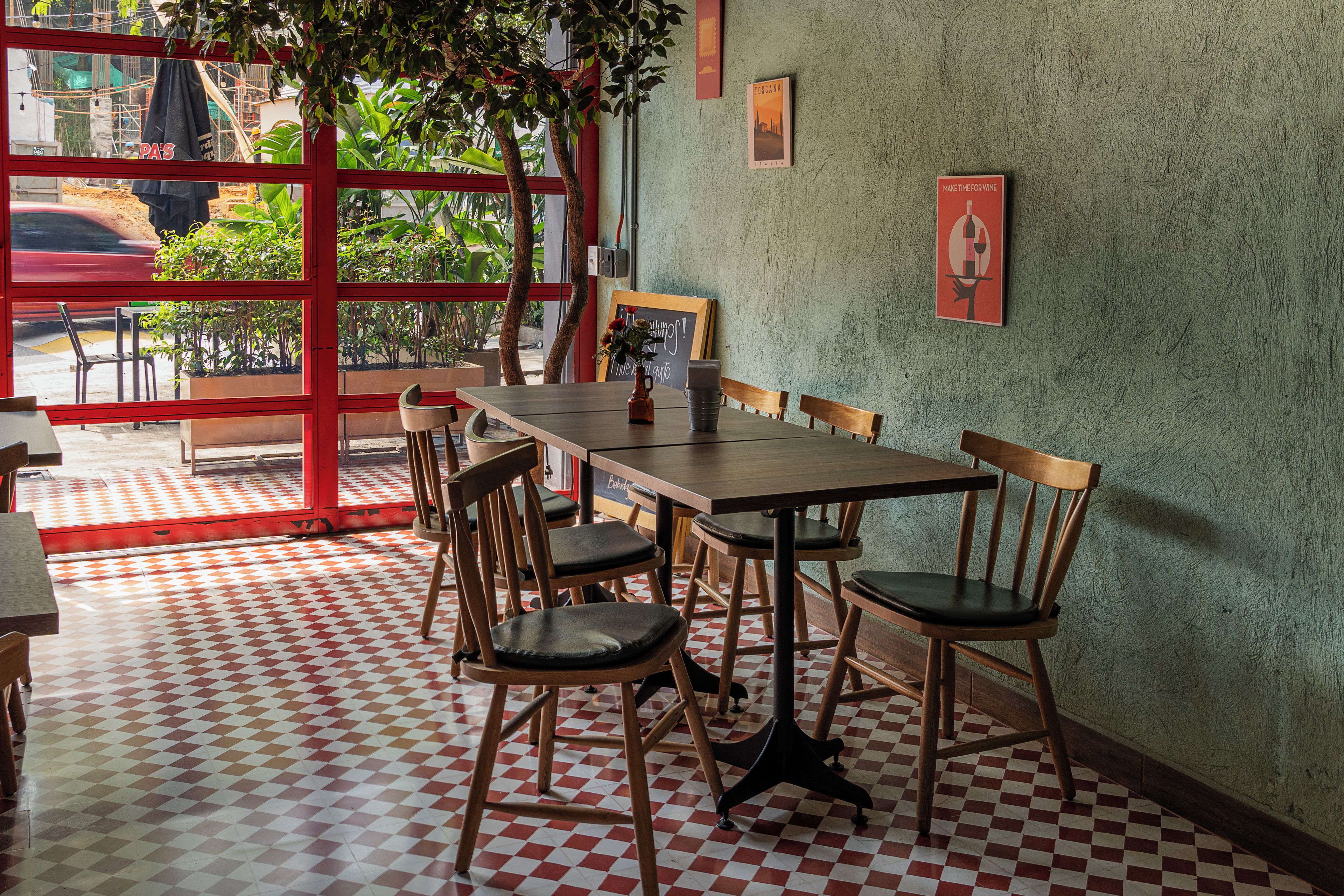 L’intérieur d’un restaurant avec un sol à carreaux, des chaises en bois, une longue table et une vue sur l’extérieur à travers une fenêtre aux montants rouges.