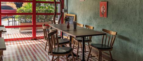 L’intérieur d’un restaurant avec un sol à carreaux, des chaises en bois, une longue table et une vue sur l’extérieur à travers une fenêtre aux montants rouges.