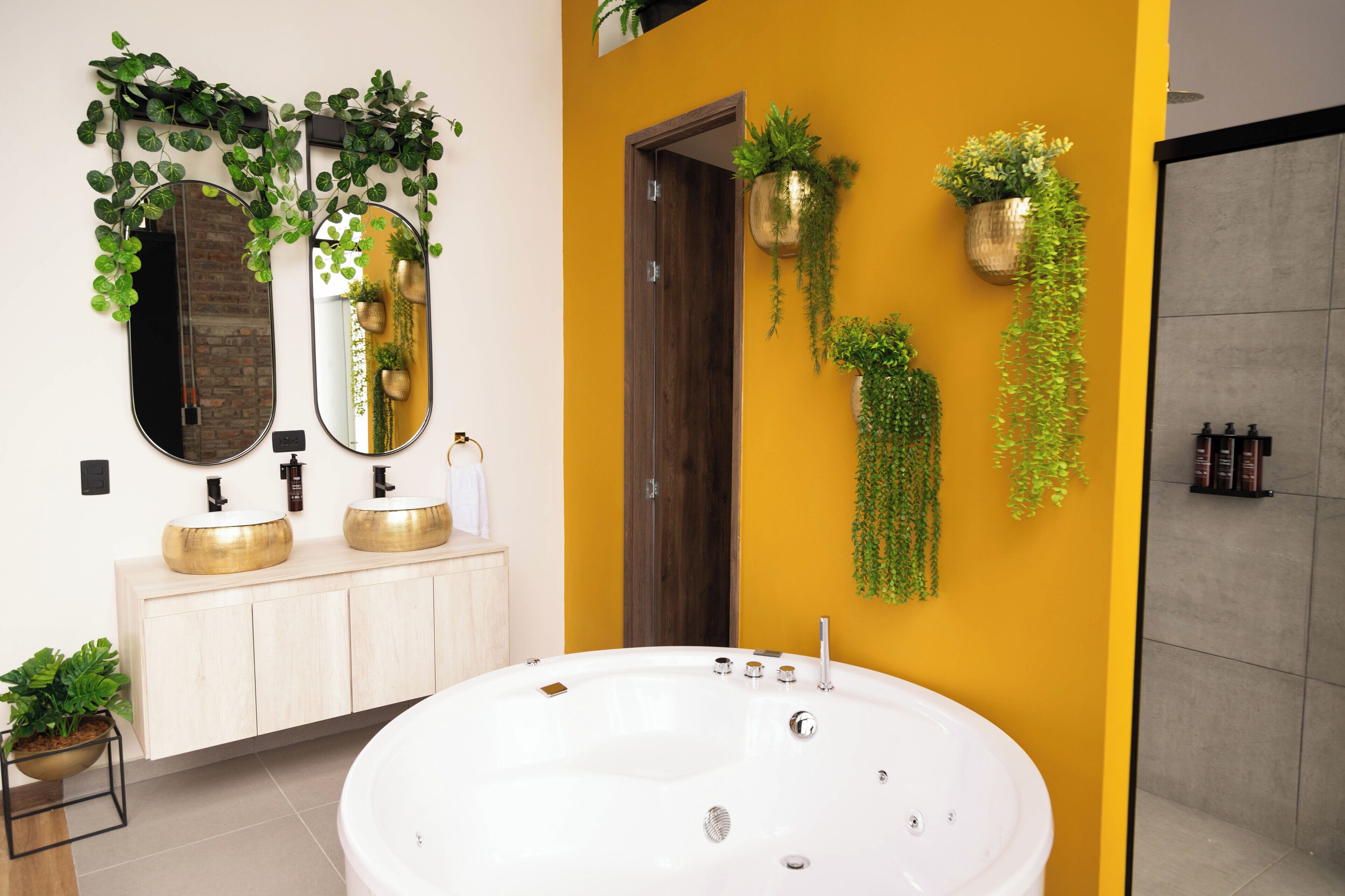 A modern bathroom with a large white bathtub, two gold sinks, and mirrors above. There are potted plants on the wall and a wooden door.