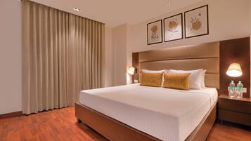 A hotel room with a large bed, wooden flooring, a headboard, two bedside lamps, and a nightstand with water bottles.