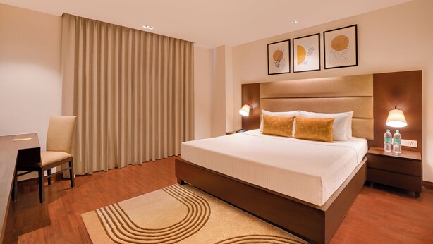 A hotel room with a large bed, a desk, a chair, and a nightstand with lamps. - Lemon Tree Hotel, Noida (Noida)