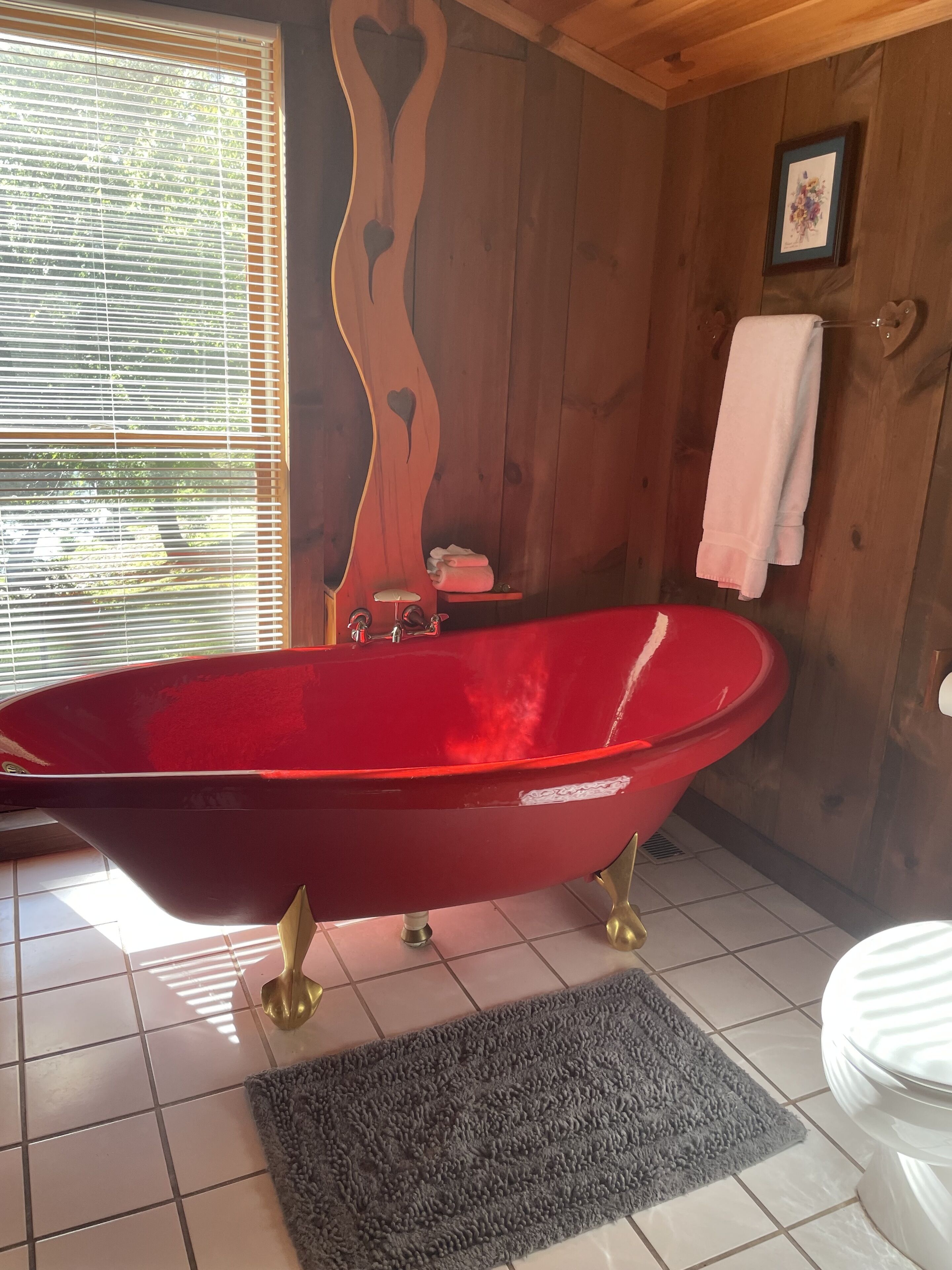 Una bañera roja tipo pata de garra en un baño de madera con suelo de baldosas, un inodoro blanco y una ventana con persianas.