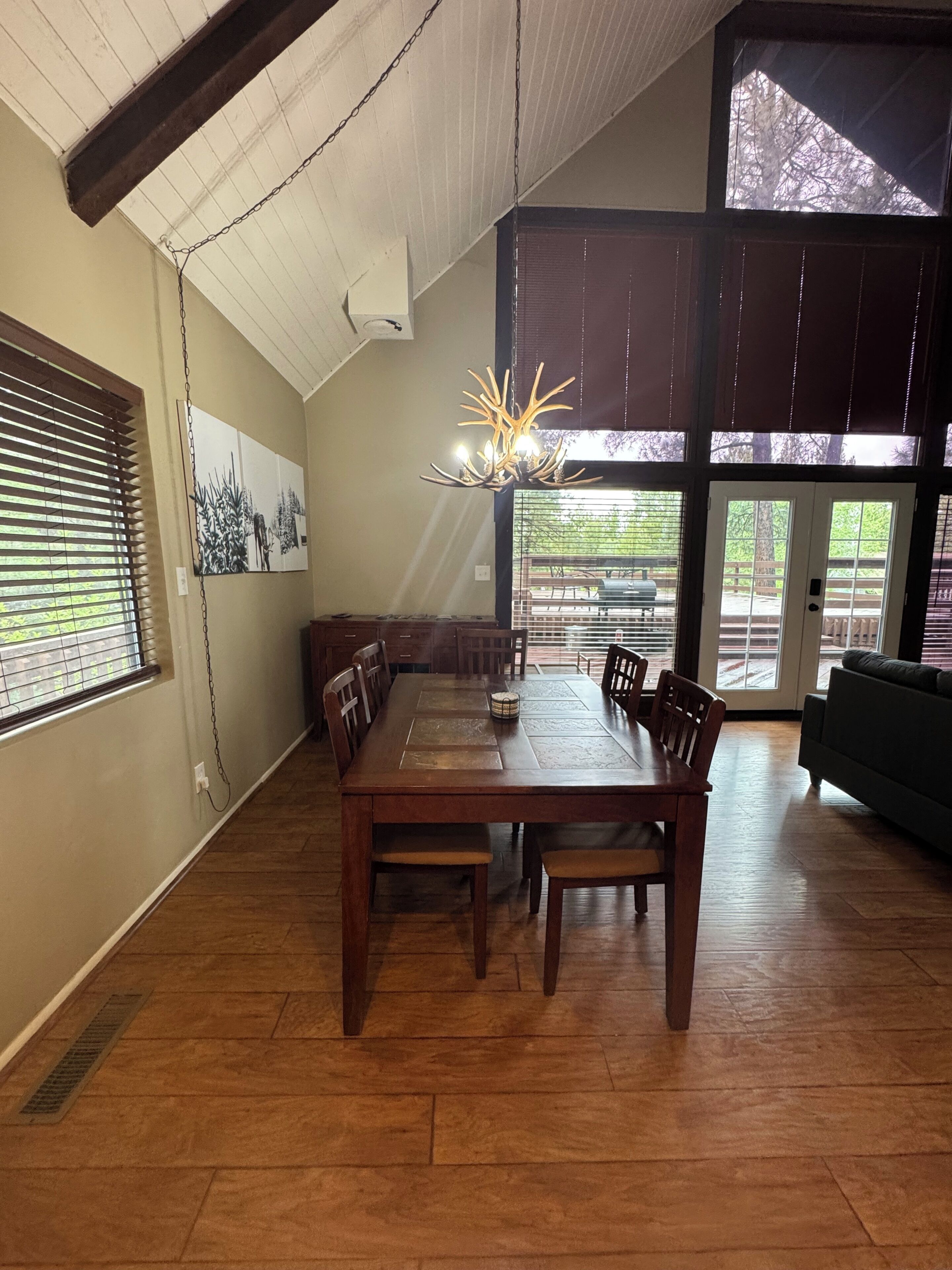 Un espace repas avec une table et des chaises en bois, un lustre et une vue sur l’extérieur grâce à de grandes fenêtres.