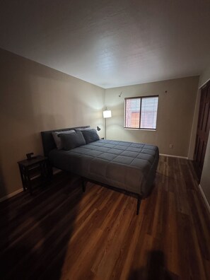 Une chambre à coucher avec un lit, une table de chevet, une lampe sur pied et une fenêtre avec des stores.