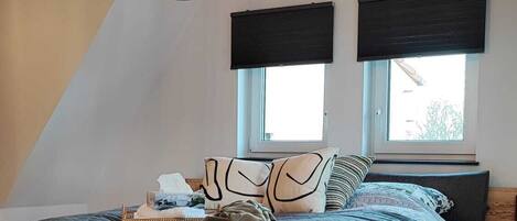A bedroom with a sloped ceiling, a bed with a grey quilt, a hanging light fixture, and two windows with blinds.
