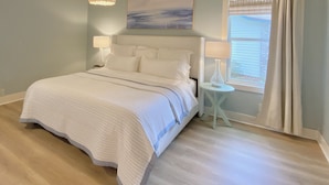 A bedroom with a large bed, two bedside tables, a chandelier, a window with curtains, and a painting on the wall. - Beautiful beach cottage 1/2 mile to the Sandy Shore! (Fernandina Beach)