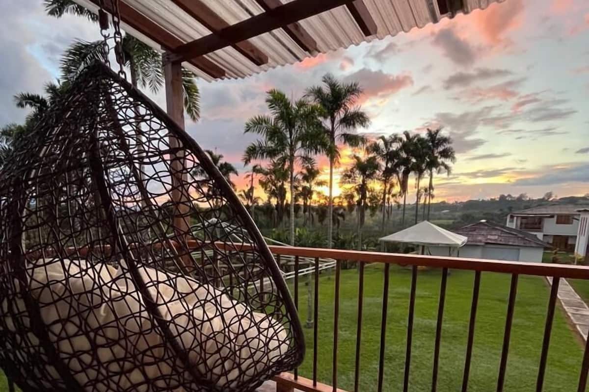 A hanging chair on a balcony with a view of palm trees and a sunset.