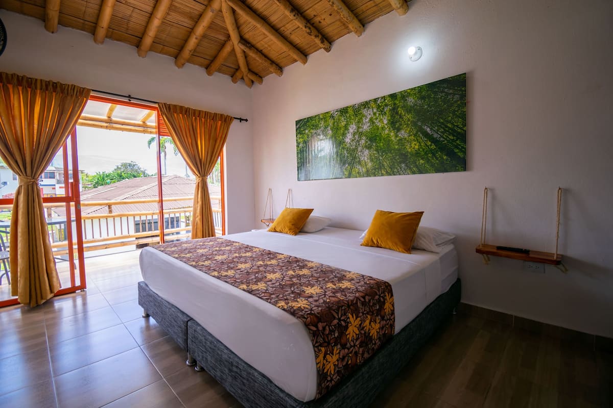 A bedroom with a large bed, a balcony with curtains, a painting on the wall, and a wooden ceiling.