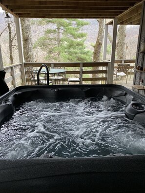 A hot tub with a wooden deck and chairs, surrounded by trees.