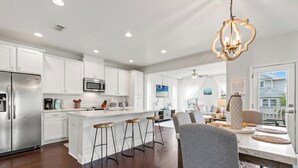 A modern kitchen with white cabinets, a stainless steel refrigerator, a microwave, and a large island with bar stools. - 30A Beach House - Whatcha Dune by PHG (Panama City Beach)