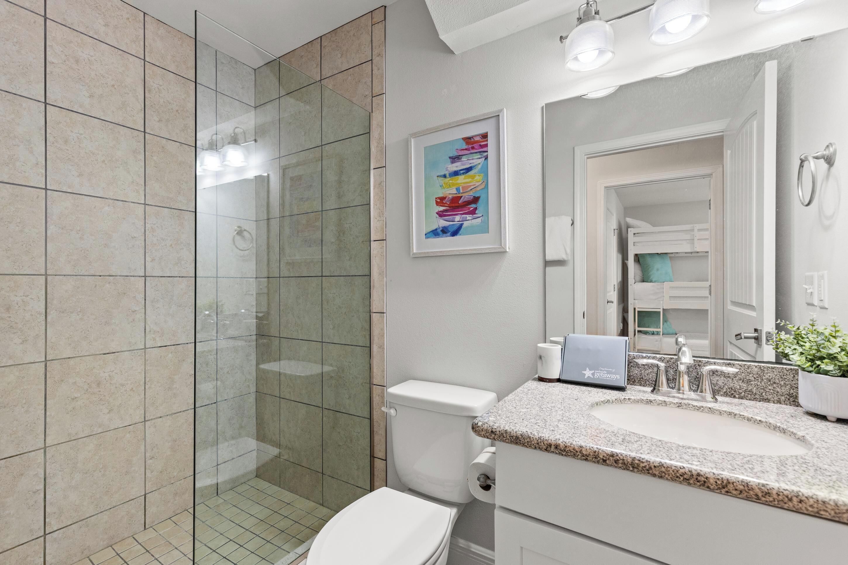 A modern bathroom with a glass shower enclosure, a white toilet, a granite countertop sink, and a framed artwork on the wall.