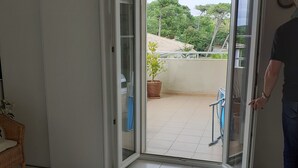 Una camera da letto con un letto, una sedia in vimini, un tavolino e un balcone con una pianta in vaso.