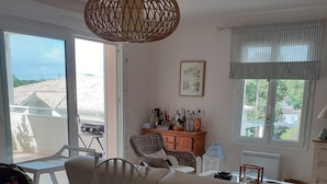 Un soggiorno moderno con un tavolo da pranzo in vetro, sedie bianche, un divano bianco, una poltrona in vimini, una lampada a sospensione in vimini, un armadietto in legno con bottiglie, un quadro incorniciato e una finestra con vista su edifici e alberi.