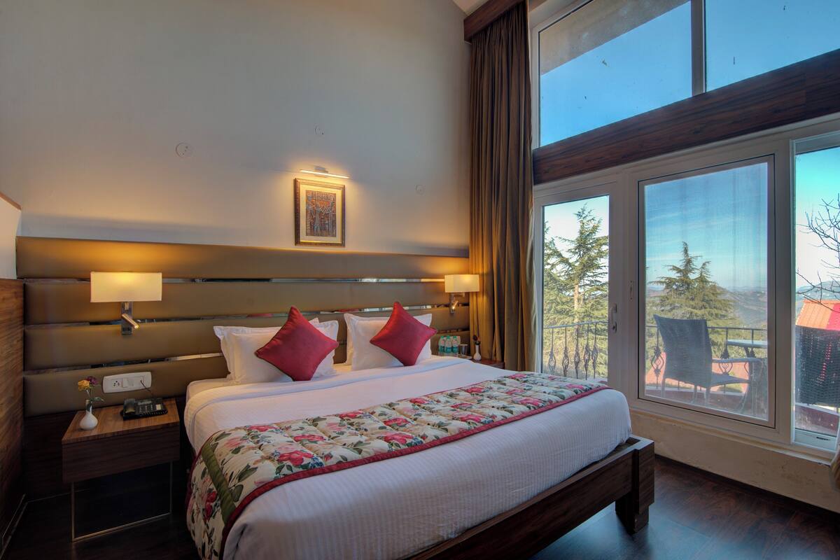 A hotel room with a large bed, a nightstand with a lamp, a window with a view of trees, and a wooden headboard.