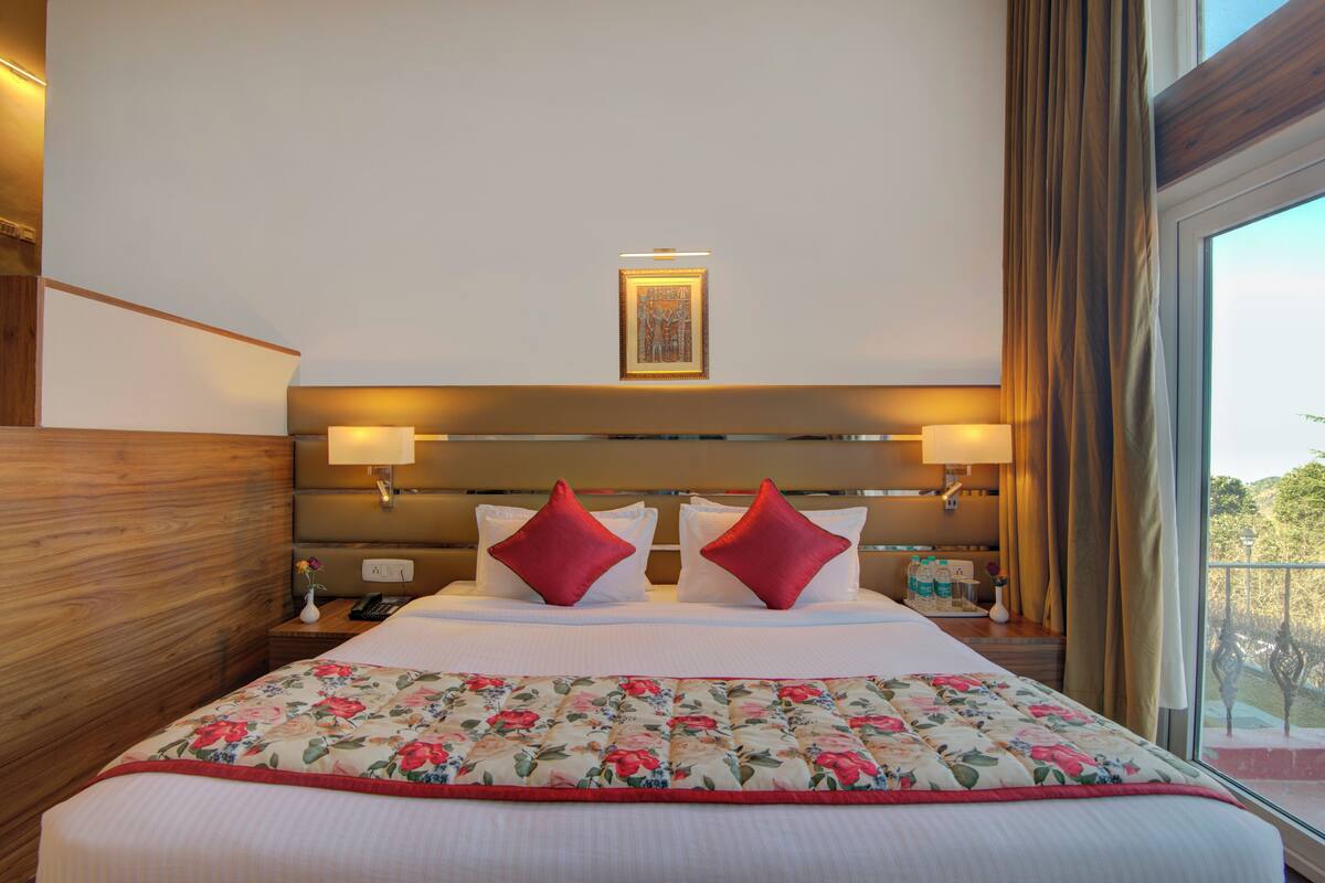 A hotel room with a large bed, two red pillows, a floral bedspread, a nightstand with a phone, and a window with curtains.