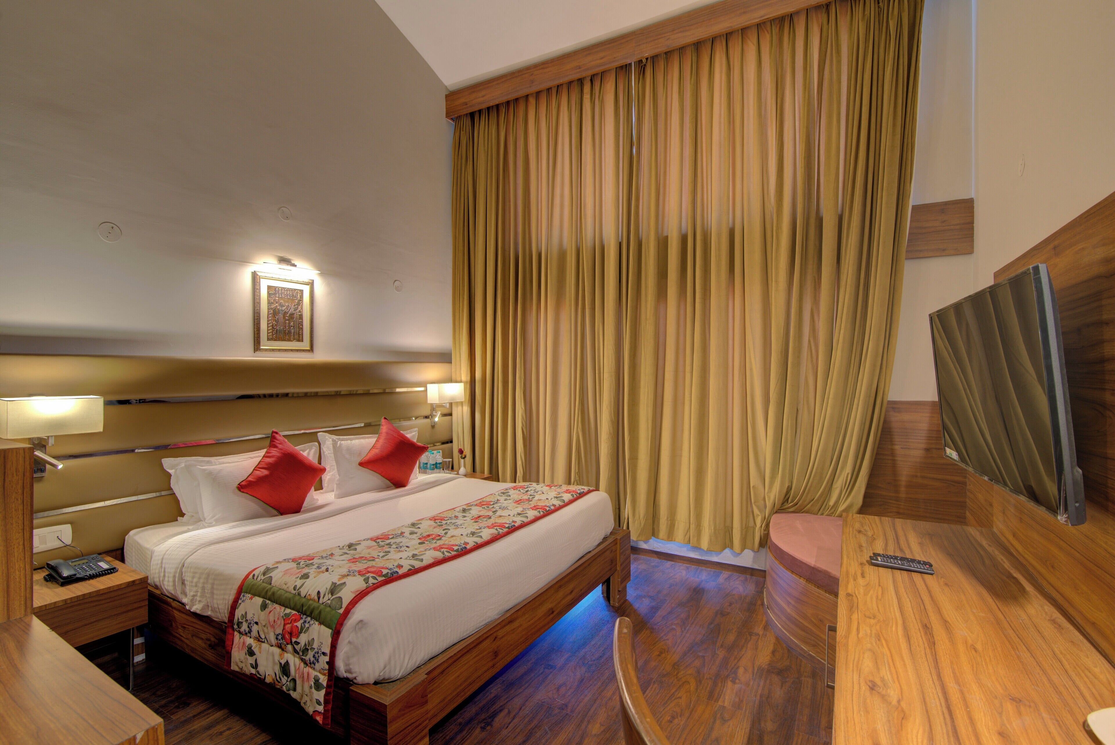 A hotel room with a large bed, wooden headboard, a desk, a chair, a TV, and a large window with curtains.