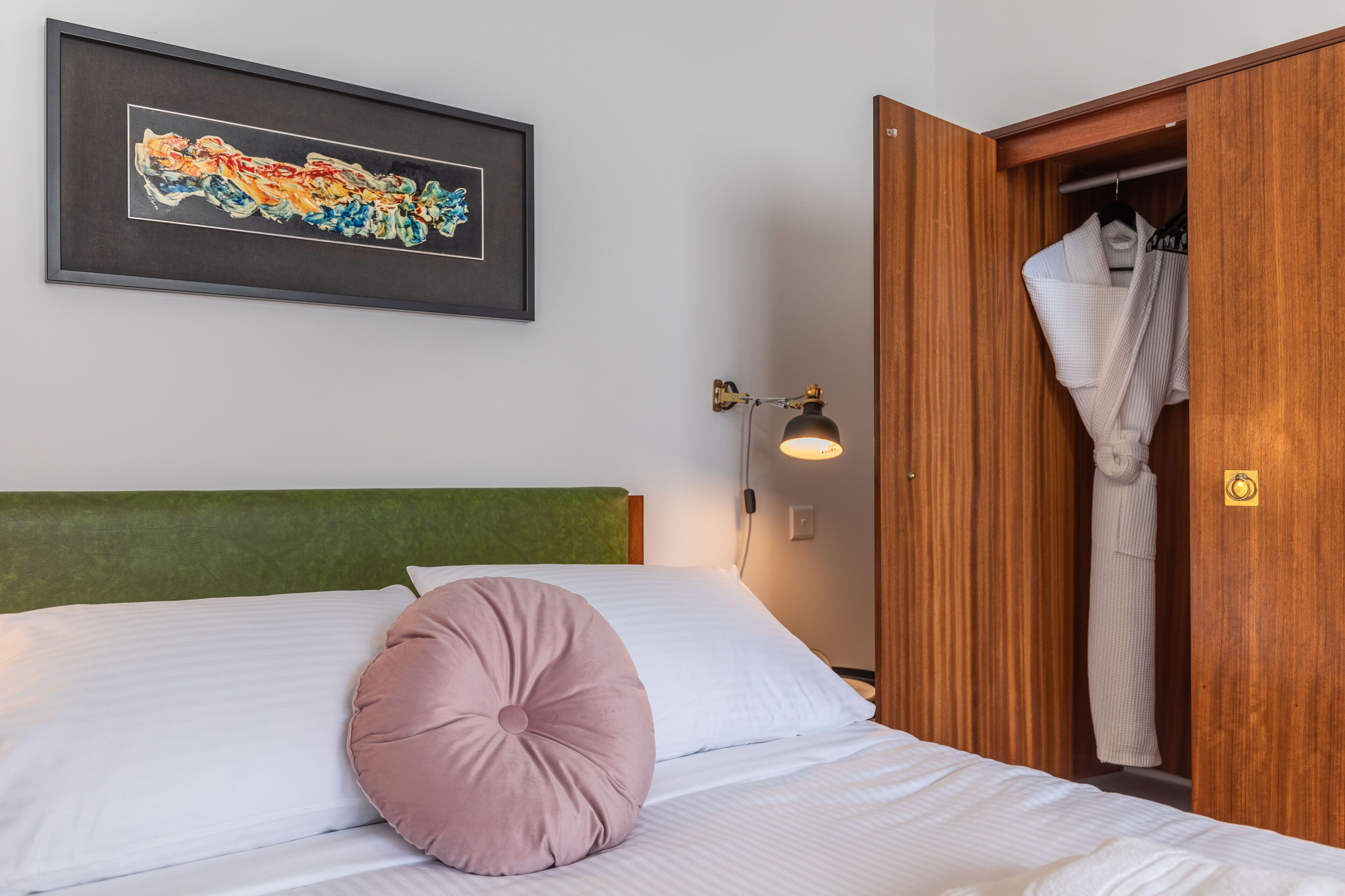 Un lit bien fait, avec des draps blancs et un seul oreiller rose, un tableau abstrait encadré accroché au mur et une armoire en bois dans laquelle est suspendu un peignoir.