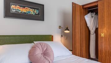 Un lit bien fait, avec des draps blancs et un seul oreiller rose, un tableau abstrait encadré accroché au mur et une armoire en bois dans laquelle est suspendu un peignoir.