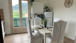A dining area with a table set for two, white chairs, a white refrigerator, a potted plant, and a view of green hills through a glass door. - Charming house in the Provencal Baronies (L'Épine)