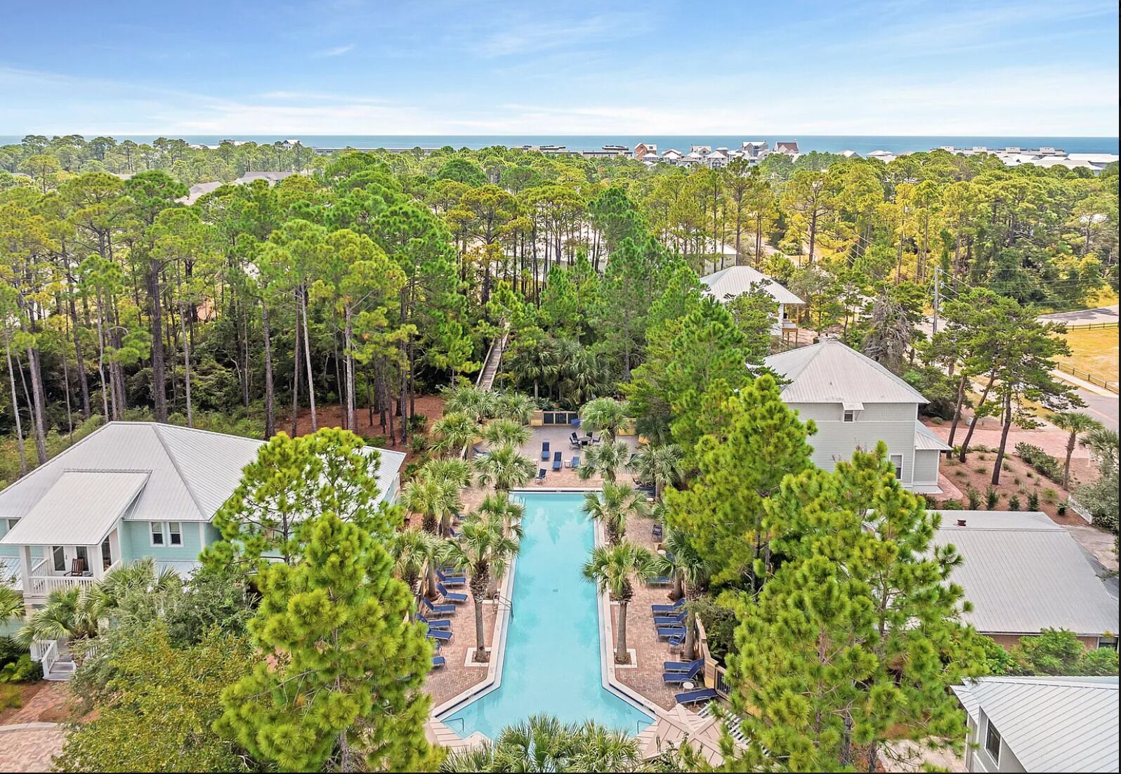 A resort with a central swimming pool surrounded by lounge chairs, surrounded by dense pine trees, and a view of the ocean.