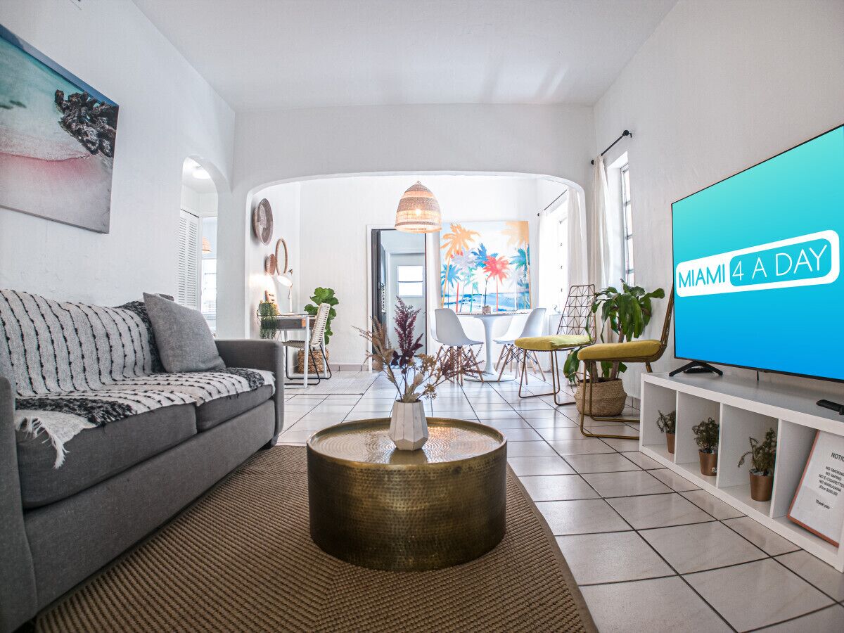 A modern living room with a grey sofa, a coffee table, and a TV displaying 'MIAMI 4 A DAY'.