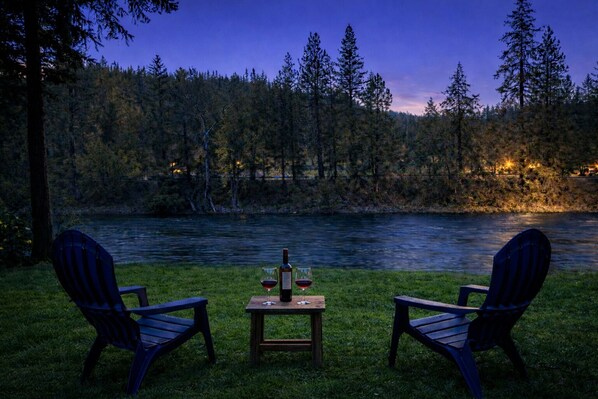Deux fauteuils Adirondack sont placés face à une rivière, avec une table entre eux, une bouteille et un verre de vin rouge.