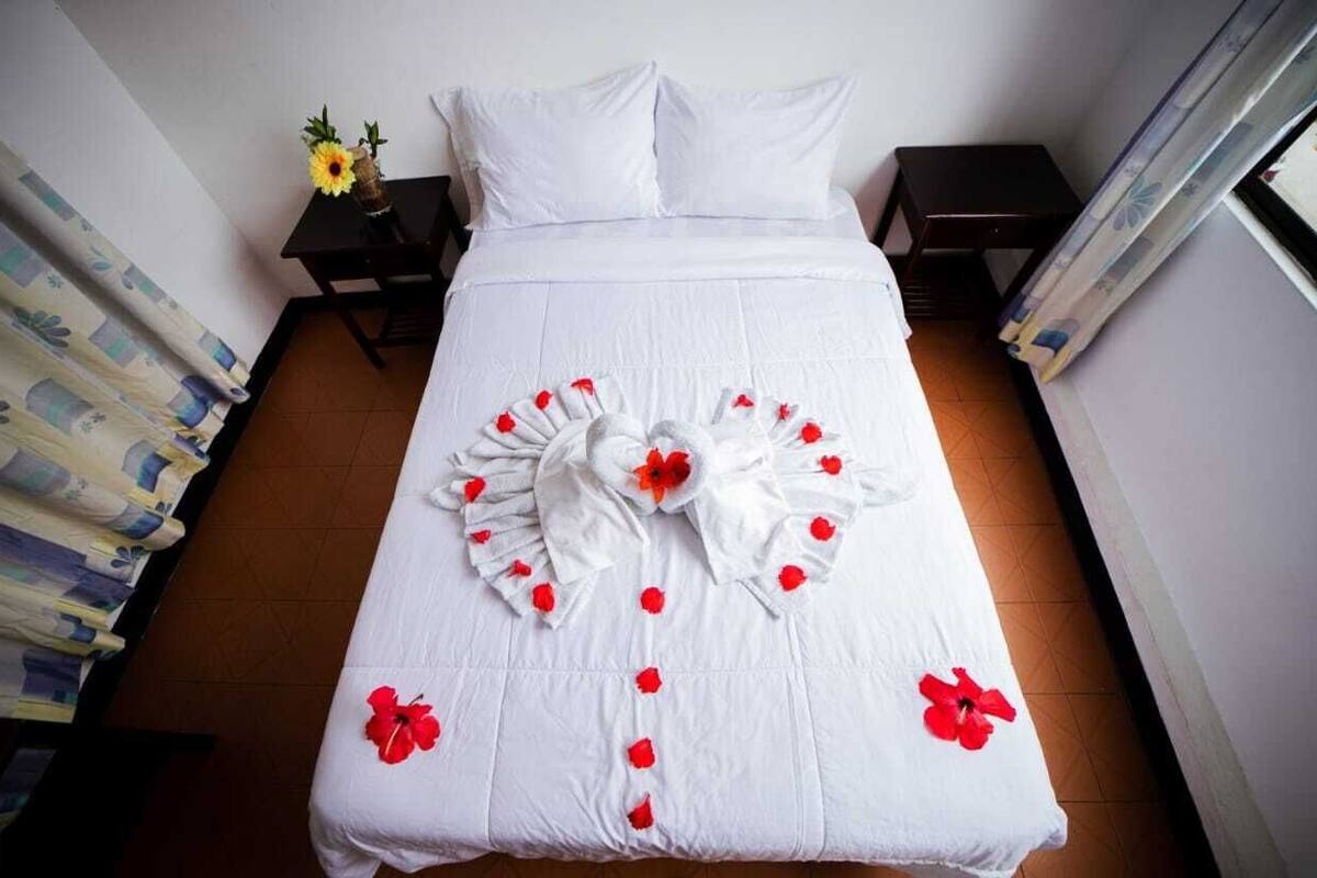 A neatly made bed with a white bedspread, red rose petals arranged in a heart shape, and a single red rose on the left side.