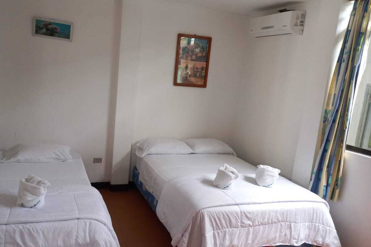 A room with two single beds, white bedding, and towels. There are two framed pictures on the walls, a window with curtains, and an air conditioning unit.