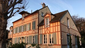 Ett traditionellt hus med korsvirkesfasad, symmetrisk utformning, stora fönster och en välskött trädgård med krukväxter och en gräsmatta.
