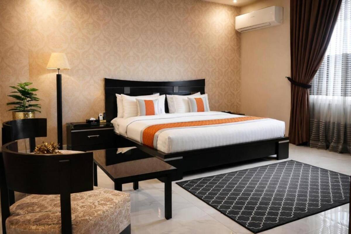 Une chambre d’hôtel avec un lit, des tables de chevet, une lampe, une plante et un tapis à motifs.