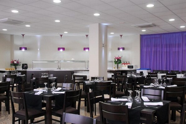 A dining area with multiple tables and chairs, a kitchen in the background, and purple curtains. - Moremar Hotel (Lloret de Mar)