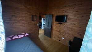Puinen huone, jossa on sänky, seinälle asennettu televisio ja ovi, joka johtaa toiseen huoneeseen.