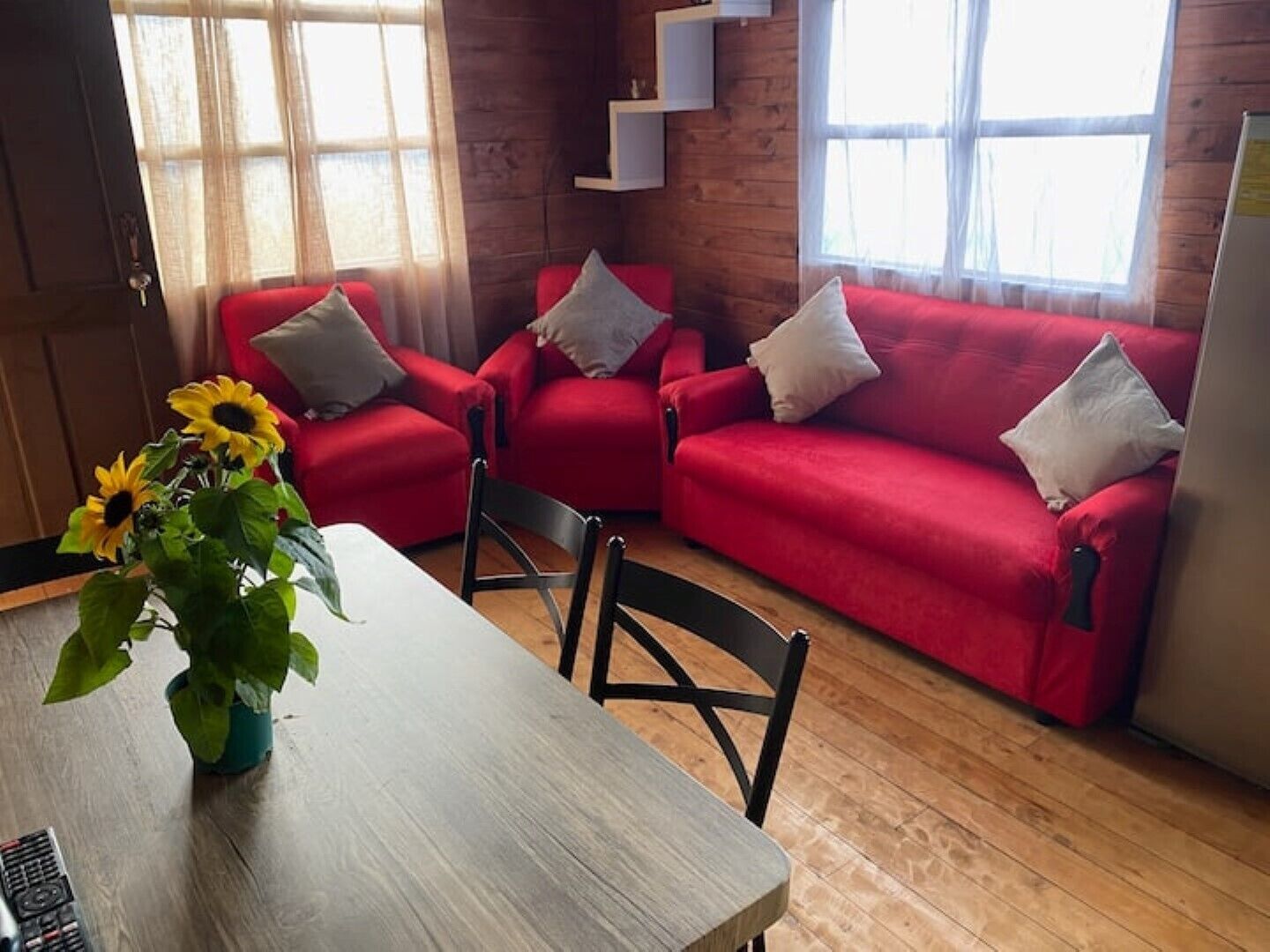 A cozy living area with a wooden floor, red sofas, a dining table with a vase of sunflowers, and a TV on a wooden shelf.