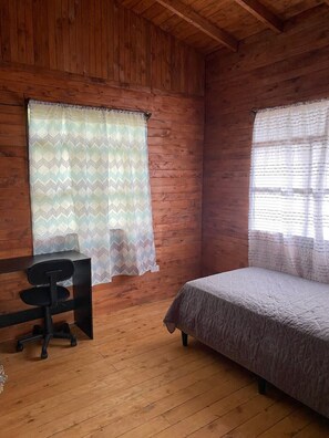 Una habitación de cabaña de madera con una cama, un escritorio y dos ventanas con cortinas estampadas.