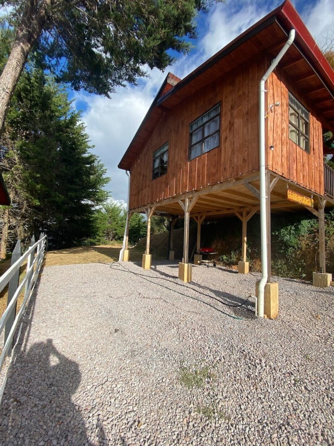 A wooden cabin with a covered porch, gravel driveway, and surrounding trees.
